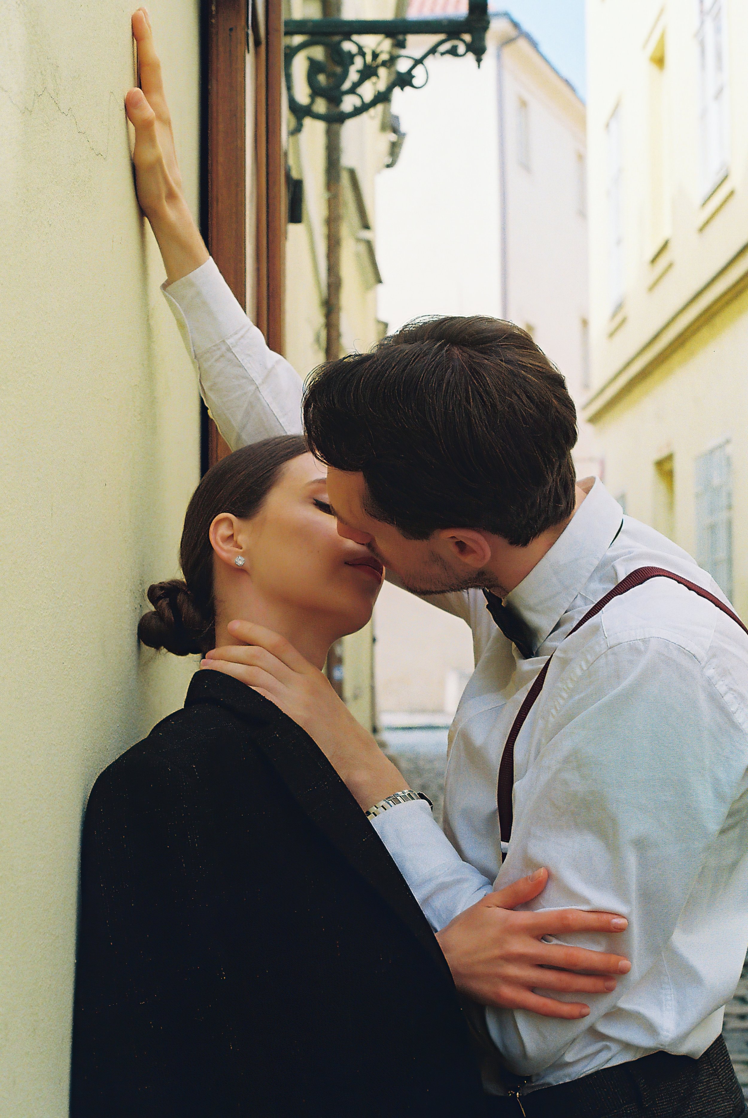 Wedding Website Design, Digital Wedding Stationery, Web Design and Branding for Wedding Professionals. A couple kissing against a wall in a narrow street, with the man wearing a white shirt and suspenders, and the woman wearing a dark coat.
